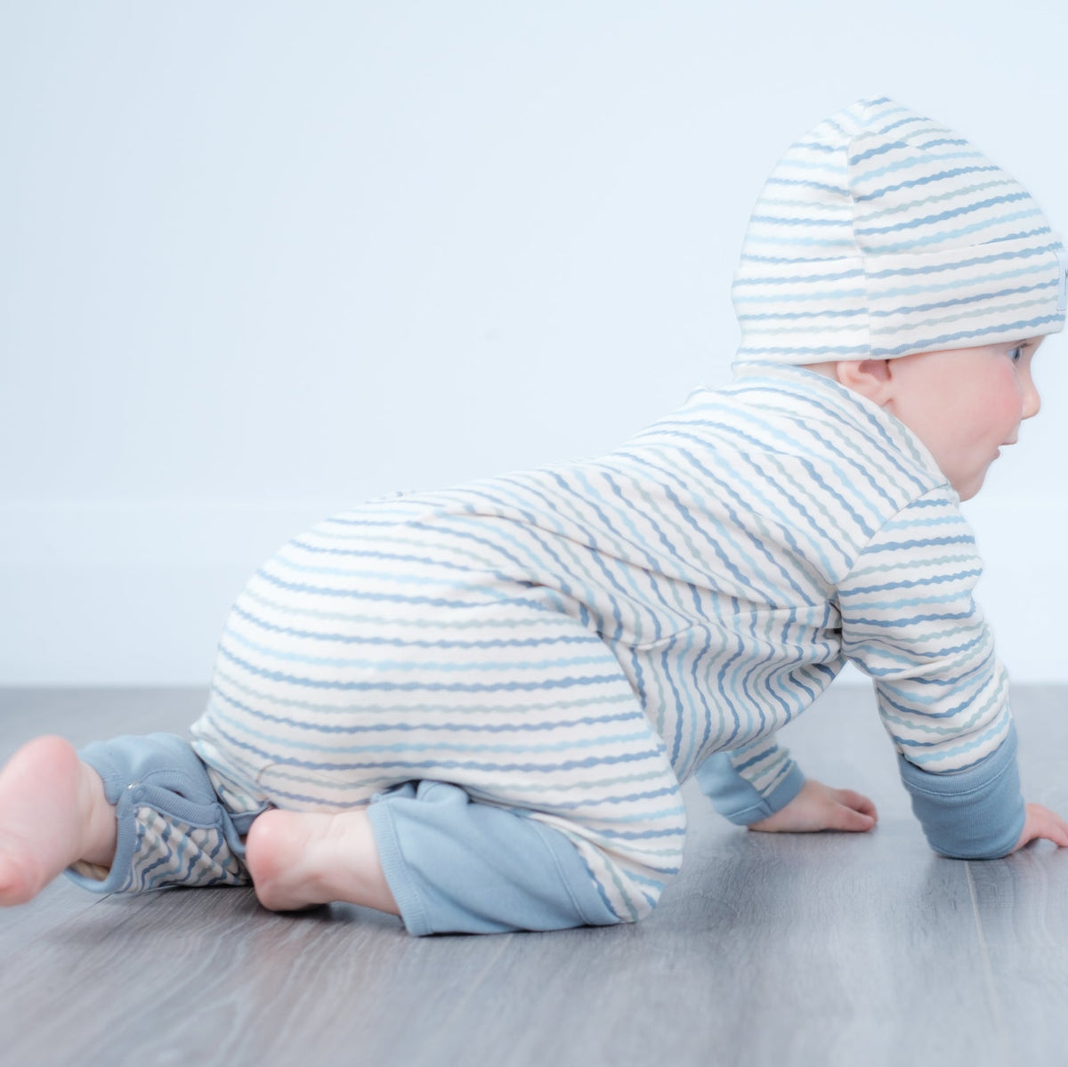 A baby crawling on a gray floor against a white wall, wearing a light blue and white striped long-sleeve romper and matching beanie hat.