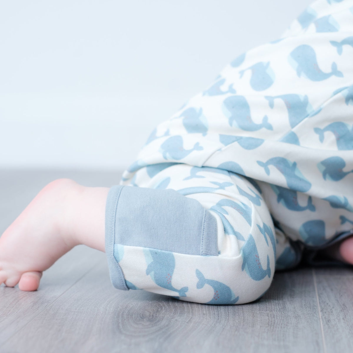 A close-up of a baby wearing a white and light blue whale-patterned one-piece outfit, crawling on a gray wood floor.