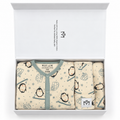 A folded baby outfit set in a white gift box, featuring a cream-colored baby grow with a sage green collar and piping, printed with penguins and snow motifs, alongside matching folded bib and hat.