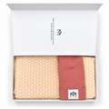 An open white gift box containing folded baby apparel: a beige patterned muslin cloth on the left, a light pink folded hat in the center, and another pink bandana bib on the right. The inside lid shows the brand name and logo printed in black.