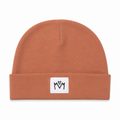 A terracotta orange knit beanie hat with a folded cuff, featuring a small white square patch on the front with a black embroidered 'M' logo.